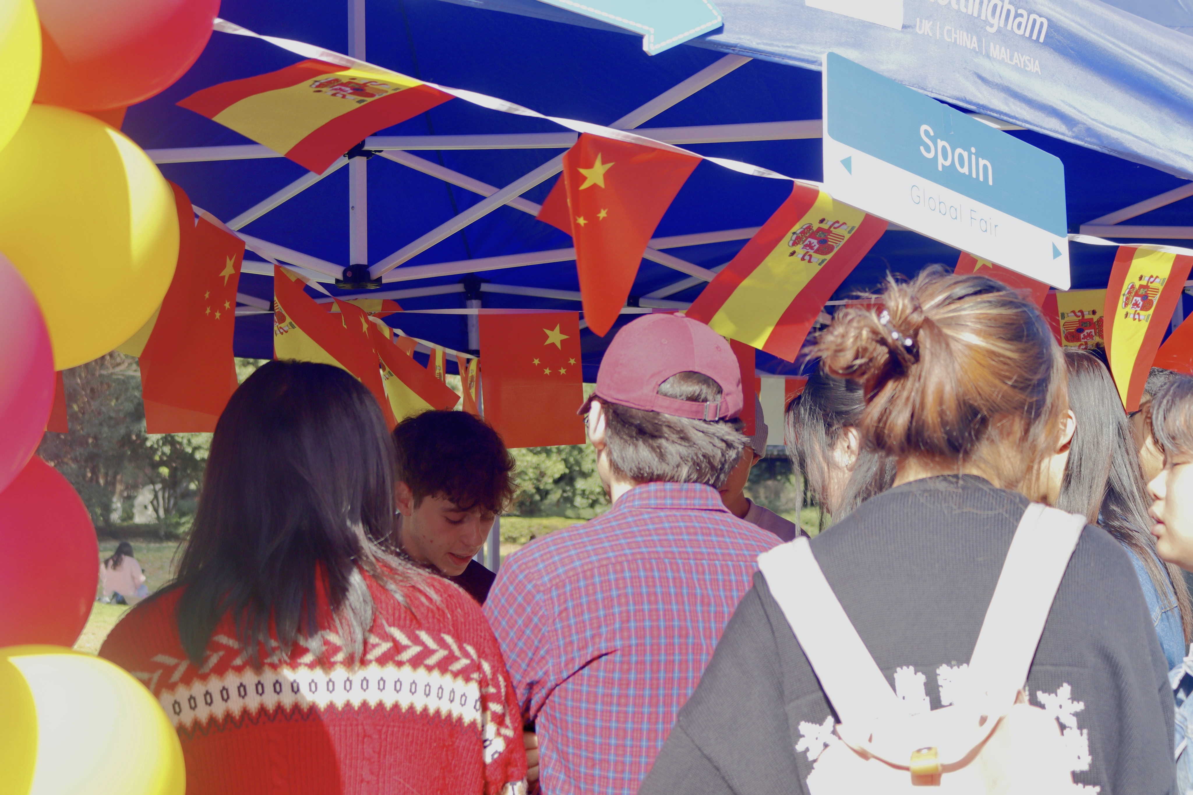 Photograph during the Global Fair at the UNNC. A group of students face the booth for Spain while Saúl explains the rules of a Spanish vocabulary game.